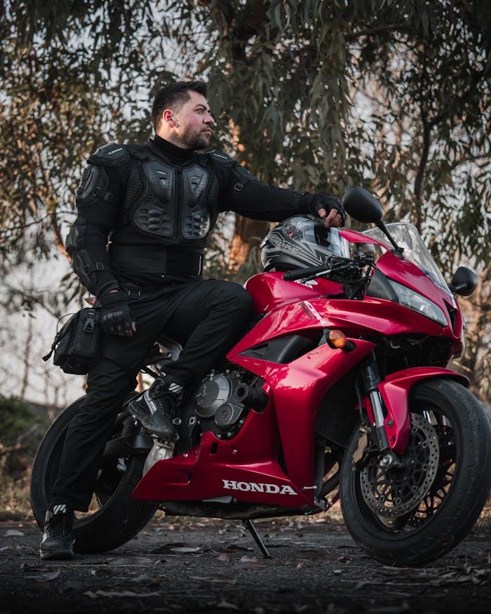Man in protective gear leaning on a red Honda motorcycle outdoors.